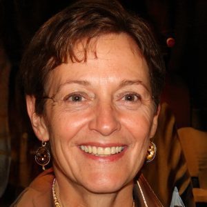 A middle-aged woman with short brown hair, wearing earrings and a brown jacket, smiles warmly at the camera. Set against a dark, blurred background, her confident expression could grace a 2024 Sale Page or Training Scale guide.