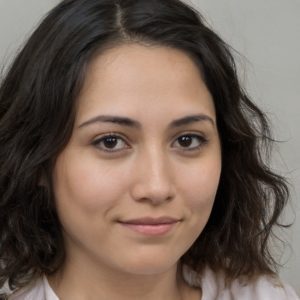 A young woman with wavy dark brown hair and fair skin looks at the camera with a neutral expression, wearing a light-colored top against a plain 2024 Training Scale background.