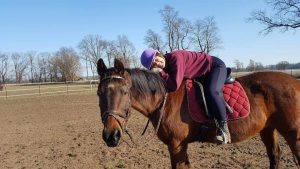 Marcee, a member of Strides, wears a purple helmet and maroon sweater while hugging the neck of a brown horse with a red saddle pad in an open field on a sunny day, surrounded by trees and fences.