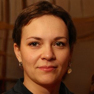A woman with short brown hair and light skin looks at the camera, wearing earrings and a dark top, with a neutral expression against a softly blurred indoor background, resembling a professional portrait for a 2024 training scale sale page.