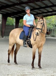 Strides member Janet, a smiling woman in a helmet, light blue shirt, and riding boots, sits on a light brown horse with a dark mane and tail in an indoor riding arena.
