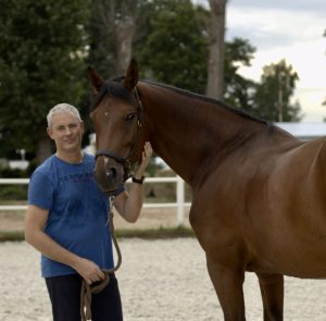 Strides member Michal, a man with short gray hair in a blue T-shirt, stands next to a brown horse, holding its lead and petting its face in an outdoor riding arena with trees and a fence in the background.