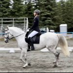 A person in a riding helmet and jacket participates in an equestrian event, riding a white horse in an outdoor arena with trees and empty bleachers in the background. Another rider on a brown horse is visible in the distance.