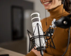 A woman wearing a brown turtleneck and headphones smiles while speaking into a white 3.0 V2 microphone mounted on a stand, suggesting she is recording a podcast or doing a voiceover.
