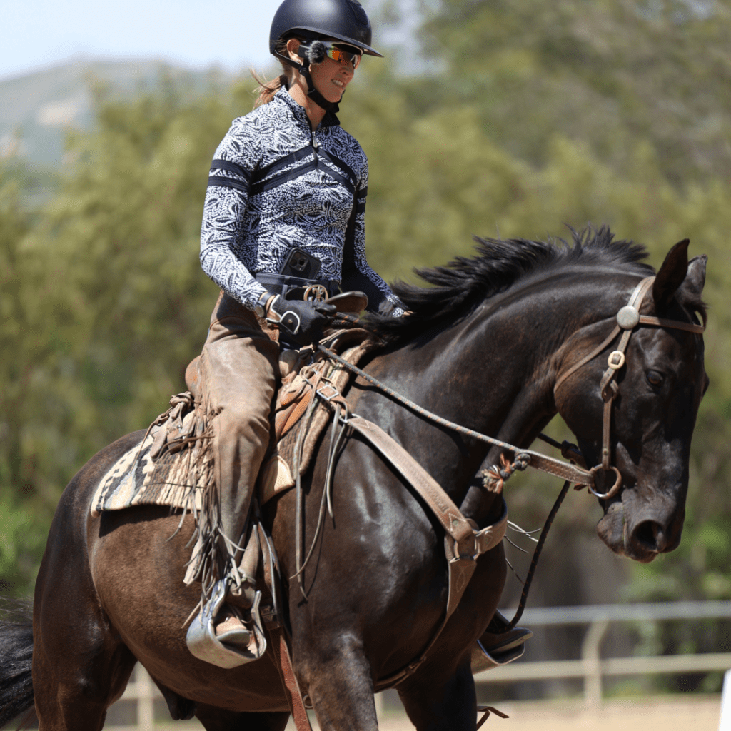 A person wearing a helmet, sunglasses, and patterned long-sleeve shirt rides a dark brown horse outdoors on a sunny day, enjoying the 30 Days Challenge with trees and a fence in the background.