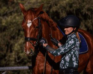 A woman in a dark helmet and patterned shirt stands beside a chestnut horse, holding its bridle and smiling, ready for a round of fitness riding among the trees in the background.