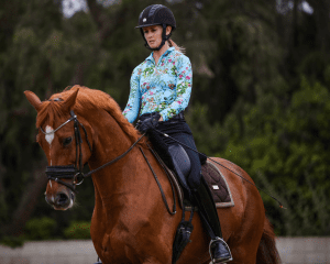 A woman wearing a black helmet and a floral long-sleeve shirt rides a chestnut horse with a white blaze on its face, outdoors amid trees and greenery—an inspiring moment on her 30 Days to Round journey.