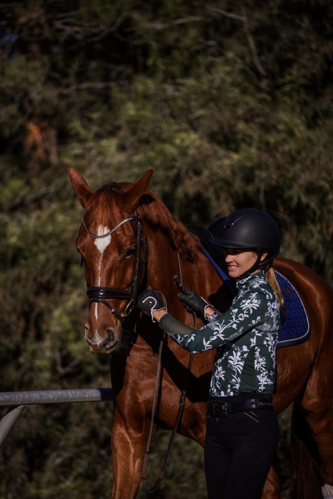 A woman wearing a helmet and a patterned shirt stands beside a brown horse with a blue saddle, smiling and patting its neck during her 30 Days to Round journey. Trees and lush greenery form the scenic background.