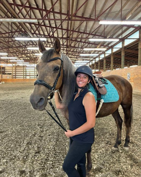 A smiling woman in a black helmet and riding attire stands next to a brown horse with a teal saddle pad inside an indoor riding arena with a high ceiling and exposed beams.