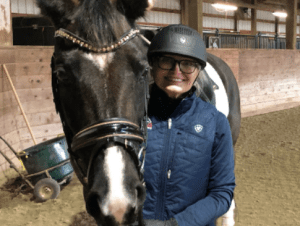 A person wearing glasses, a helmet, and a blue vest stands next to a black and white horse inside an indoor riding arena with wooden walls, ready for the Flying Changes Webinar.
