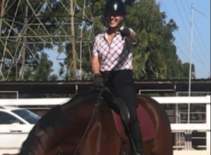A person wearing a helmet and patterned shirt is smiling and pointing while sitting on a dark brown horse in an outdoor riding arena, perhaps inspired by tips from the Flying Changes Webinar. Trees, a fence, and a white car are visible in the background.