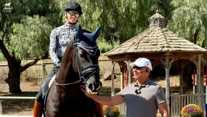 A smiling woman in riding gear sits on a dark horse, while Steffen Peters, wearing a cap and sunglasses, stands beside them, petting the horse. Behind them is a wooden gazebo and green trees—a perfect clinic recap moment.