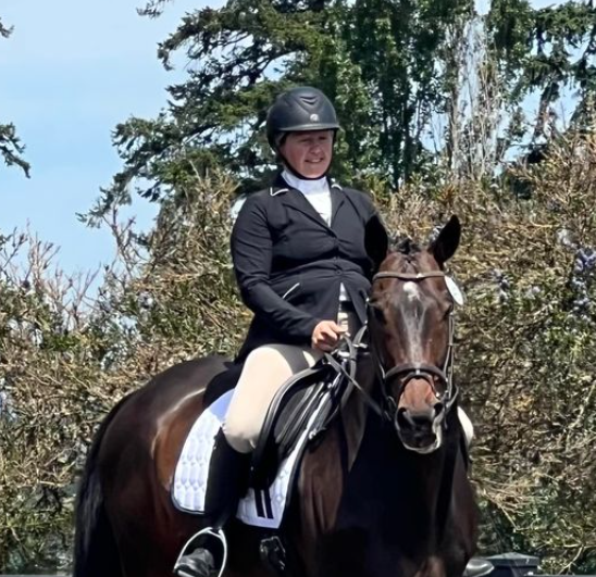 A person wearing a black helmet, black jacket, and beige riding pants is riding a dark brown horse with a white saddle pad during the Groundwork Webinar. Trees and blue sky are visible in the background.