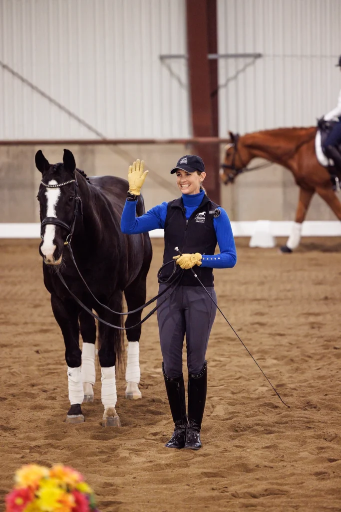 A smiling woman in riding attire stands next to a black horse with white markings, holding its reins and waving. They are indoors on a sandy arena, with another horse and rider in the background.