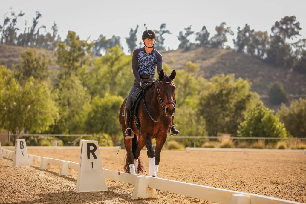 A person wearing a helmet and riding gear is riding a brown horse in an outdoor dressage arena, passing between markers labeled B X and R I, with trees and hills in the background.