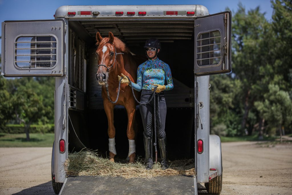 A person in riding gear stands beside a chestnut horse in a horse trailer, holding the lead and smiling. Sunshine and greenery outside suggest they've mastered groundwork essentials for safe, confident handling before the ride.