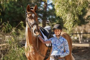 A person wearing a helmet and a blue-patterned shirt stands outdoors, smiling as they hold the reins of a brown horse with a black mane—ready for a free test ride. Trees and sunlight fill the background as they prepare to take new strides.