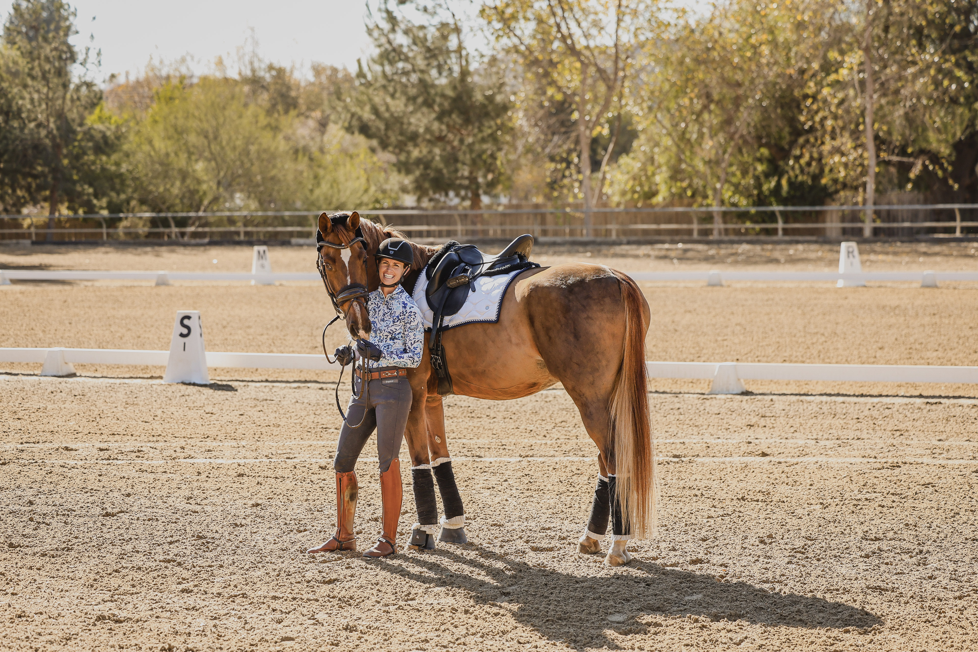 A smiling equestrian stands beside a brown horse wearing riding gear on a sandy outdoor arena, ready for a free test ride. The pair is surrounded by trees, dressage markers, and sunlit strides on a beautiful day.