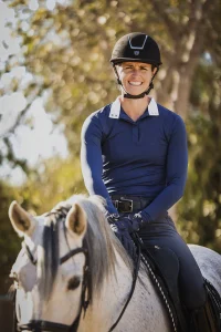 A person wearing a black helmet, blue long-sleeve shirt, and gloves is smiling while riding a white horse outdoors with sunlight and trees in the background—capturing the spirit of Strides 2024 in every stride.