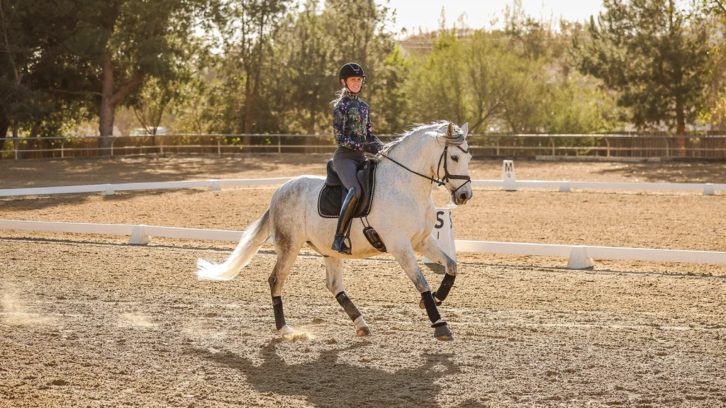 A person wearing a helmet and patterned top rides a white horse in an outdoor arena on a sunny day, performing dressage for Strides 2024, with trees and a fence in the background.