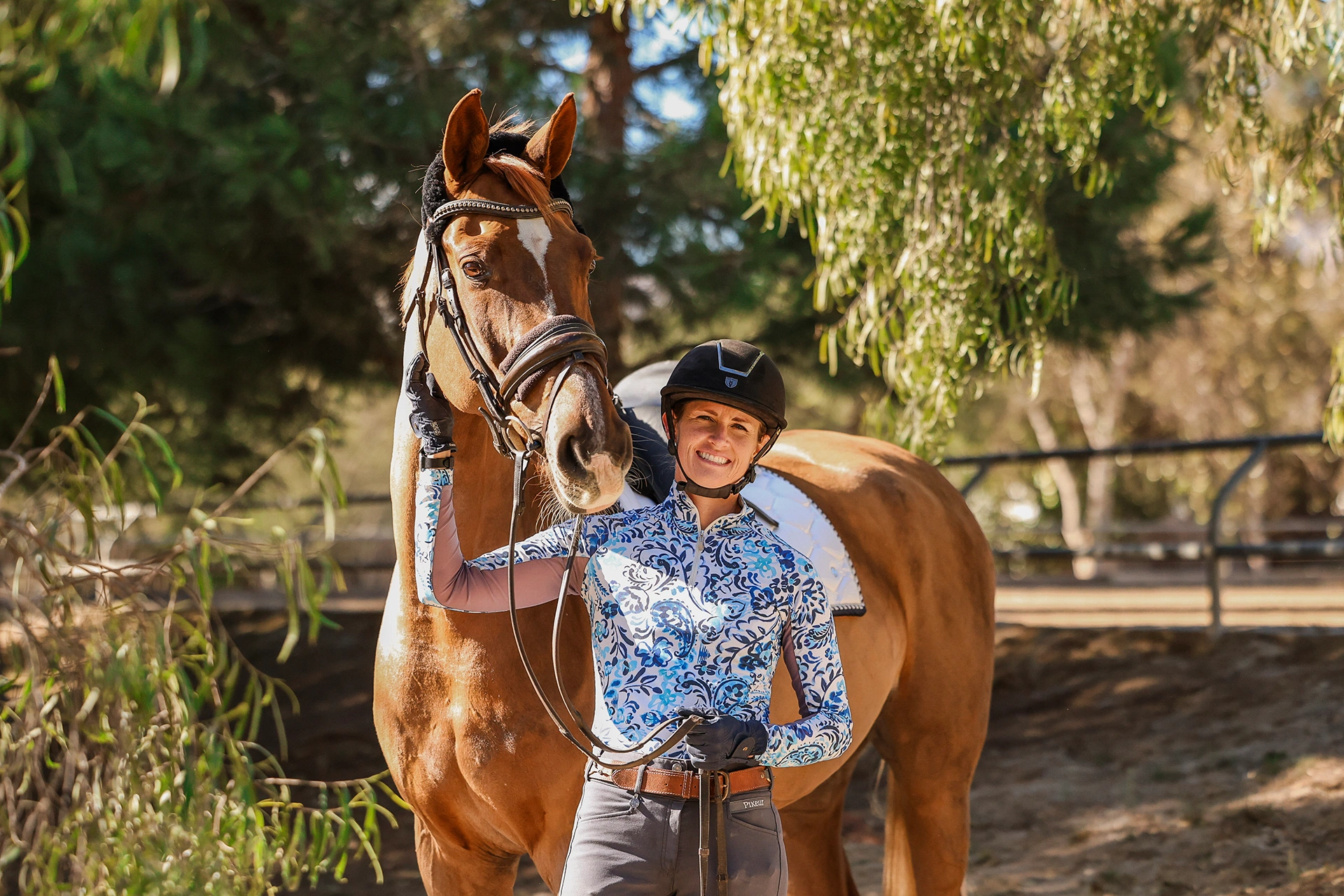 A person sporting a riding helmet, gloves, and a patterned shirt stands outdoors next to a brown horse, smiling and holding the horse’s bridle as they enjoy the sunlight—showcasing joyful Strides 2024 moments.