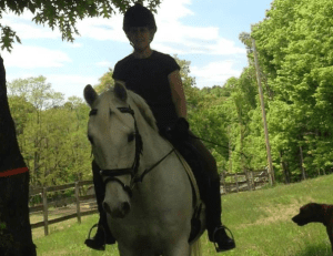 A person wearing a helmet and dark clothing rides a white horse outdoors on a grassy area, with green trees and a wooden fence in the background. Part of a brown dog is visible at the lower right—a perfect scene for the 2025 Riding Bootcamp.