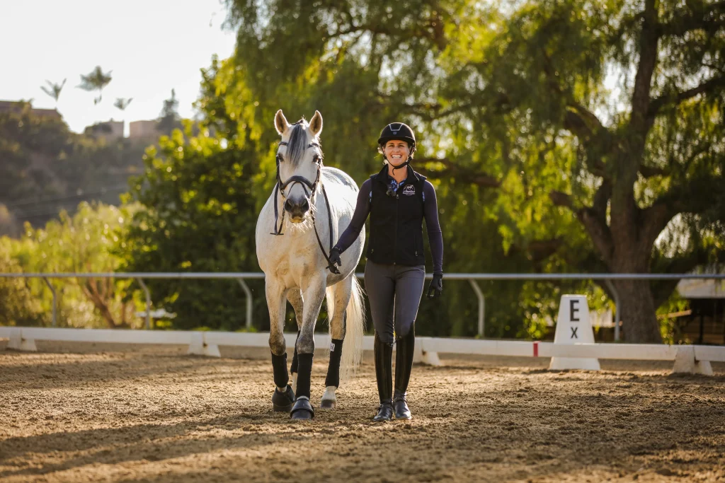 A woman in equestrian gear smiles while walking beside a white horse in an outdoor riding arena on a sunny day, with trees and fencing in the background—perfect inspiration for our Free Groundwork Webinar.