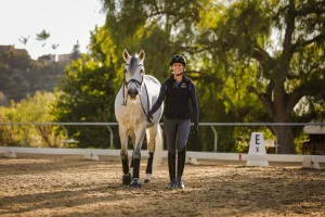 A woman in equestrian gear smiles while walking beside a white horse in an outdoor riding arena on a sunny day, with trees and fencing in the background—perfect inspiration for our Free Groundwork Webinar.