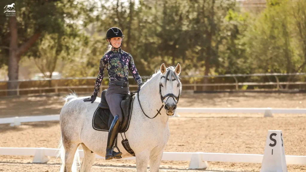 A woman wearing a helmet and a floral-patterned top sits on a white horse in an outdoor dressage arena, smiling, for our January Newsletter 2025. Trees and dressage markers are visible in the background.