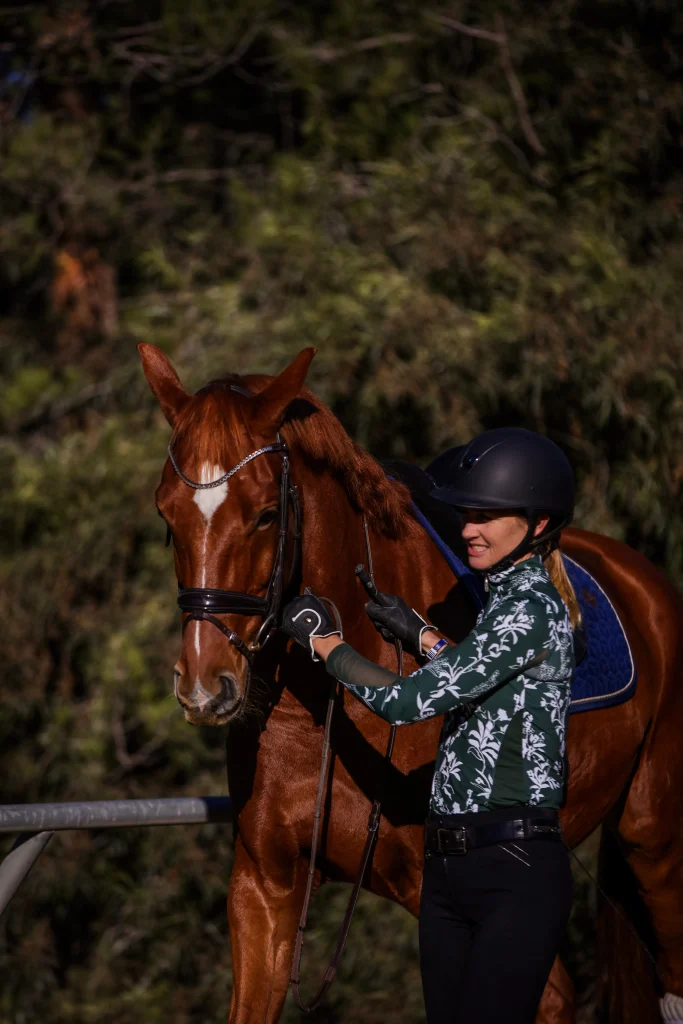 Amelia, in a riding helmet and floral shirt, stands beside a chestnut horse with a blue saddle pad, smiling and holding the reins outdoors with trees in the background—ready to master groundwork and piaffe.