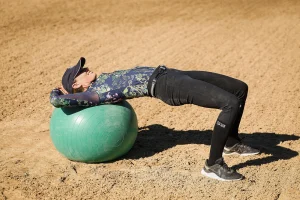 A person wearing a cap, long-sleeve floral shirt, and black pants demonstrates Rider Position by performing a bridge exercise on a green stability ball on sandy ground.