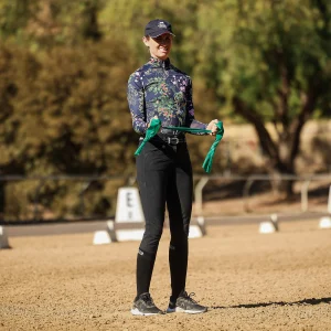 A person wearing a floral long-sleeve shirt, black pants, and a cap stands on a sandy arena holding a green ribbon, smiling—perfect for illustrating the Rider Position Blueprint. Trees and dressage markers can be seen in the background.