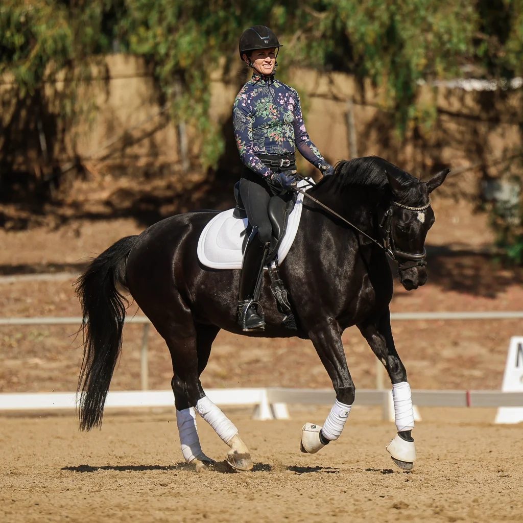 A person in a floral-patterned long-sleeve shirt, black helmet, and riding boots demonstrates the Rider Position Blueprint while riding a dark horse with white leg wraps and a saddle pad in an outdoor arena framed by trees and a fence.