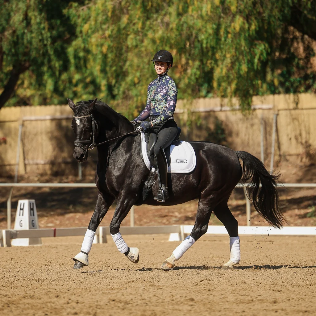 A person in a helmet and floral riding shirt demonstrates excellent rider position while riding a black horse with white gear in an outdoor sandy arena, framed by trees and a fence in the background.