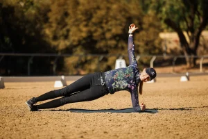 A woman wearing a floral long-sleeve shirt, black pants, and a cap is performing a side plank on a mat outdoors, demonstrating core strength essential for proper rider position in horseback riding, with trees and sand in the background.