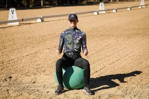 A person wearing a hat, floral long-sleeve shirt, riding pants, and boots is smiling while sitting on a green exercise ball in a sandy outdoor arena, demonstrating correct rider position. Dressage marker letters are visible in the background.