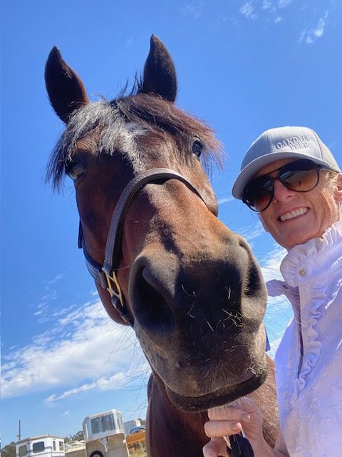 Member Becky, wearing sunglasses and a cap, smiles while standing next to a dark brown horse under a bright blue sky. Captured from a low angle, the horse’s powerful strides make it appear even larger against the scattered clouds.