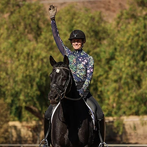 A person demonstrates the Rider Position Blueprint, wearing a helmet and floral long-sleeve shirt while riding a black horse, raising one hand to wave, with trees and sunlight in the background.