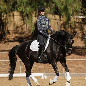 A person showcasing the Rider Position Blueprint, wearing a helmet, sunglasses, and a patterned long-sleeve shirt, rides a black horse with white leg wraps and a white saddle pad in an outdoor arena surrounded by trees.