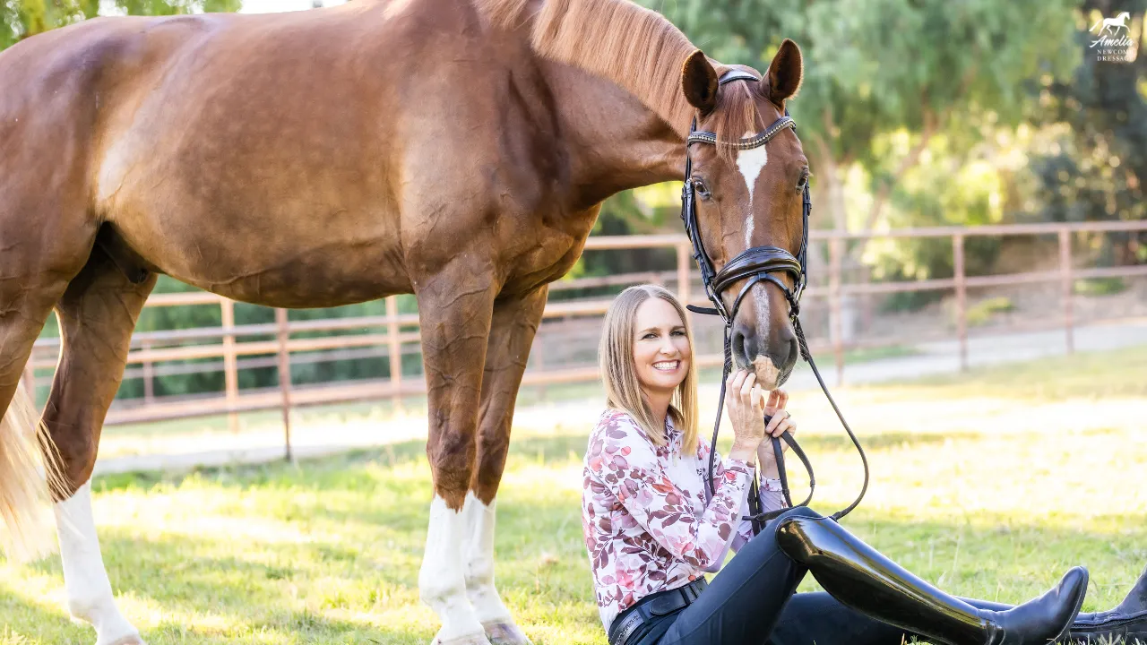 A smiling woman in riding boots sits on the grass, holding the reins of a brown horse beside her in a sunny, fenced outdoor area—a perfect Valentine scene for February 2025 with trees in the background.