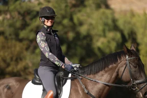 A person wearing a helmet, sunglasses, gloves, and a floral long-sleeve shirt rides a dark brown horse with a white saddle pad outdoors, surrounded by blurred green trees—perfect for listening to your favorite Confidence podcast.