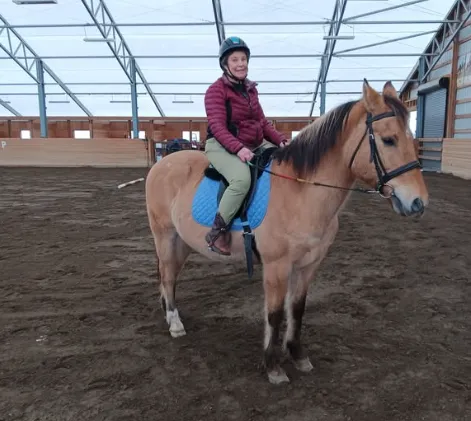 A person wearing a helmet and maroon jacket is smiling with confidence while sitting on a light brown horse with a blue saddle pad inside a spacious indoor riding arena—perfect for trying out new confidence tips.