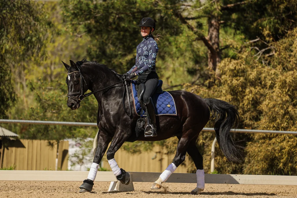 A person wearing a helmet and floral-patterned shirt rides a black horse with white leg wraps and a blue saddle pad on a sandy arena, surrounded by trees and a wooden fence—perfect for the Canter Challenge. Join now!.
