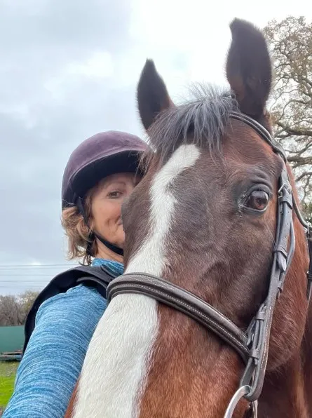 A person wearing a helmet and blue top stands close behind a brown horse with a white blaze on its face, both looking at the camera. The background shows a cloudy sky and some trees.