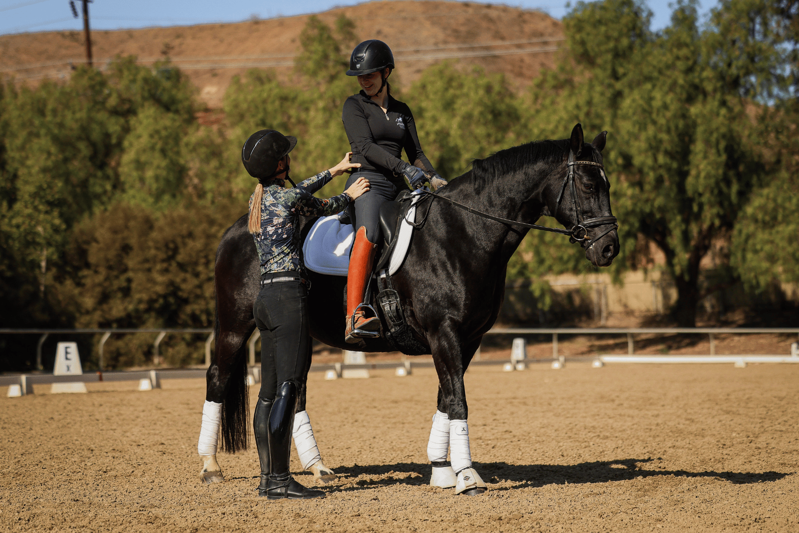 A riding instructor stands beside a woman sitting on a black horse, giving guidance in an outdoor arena as they practice Canter V1. Both wear helmets and riding gear, with trees and a hill in the background.
