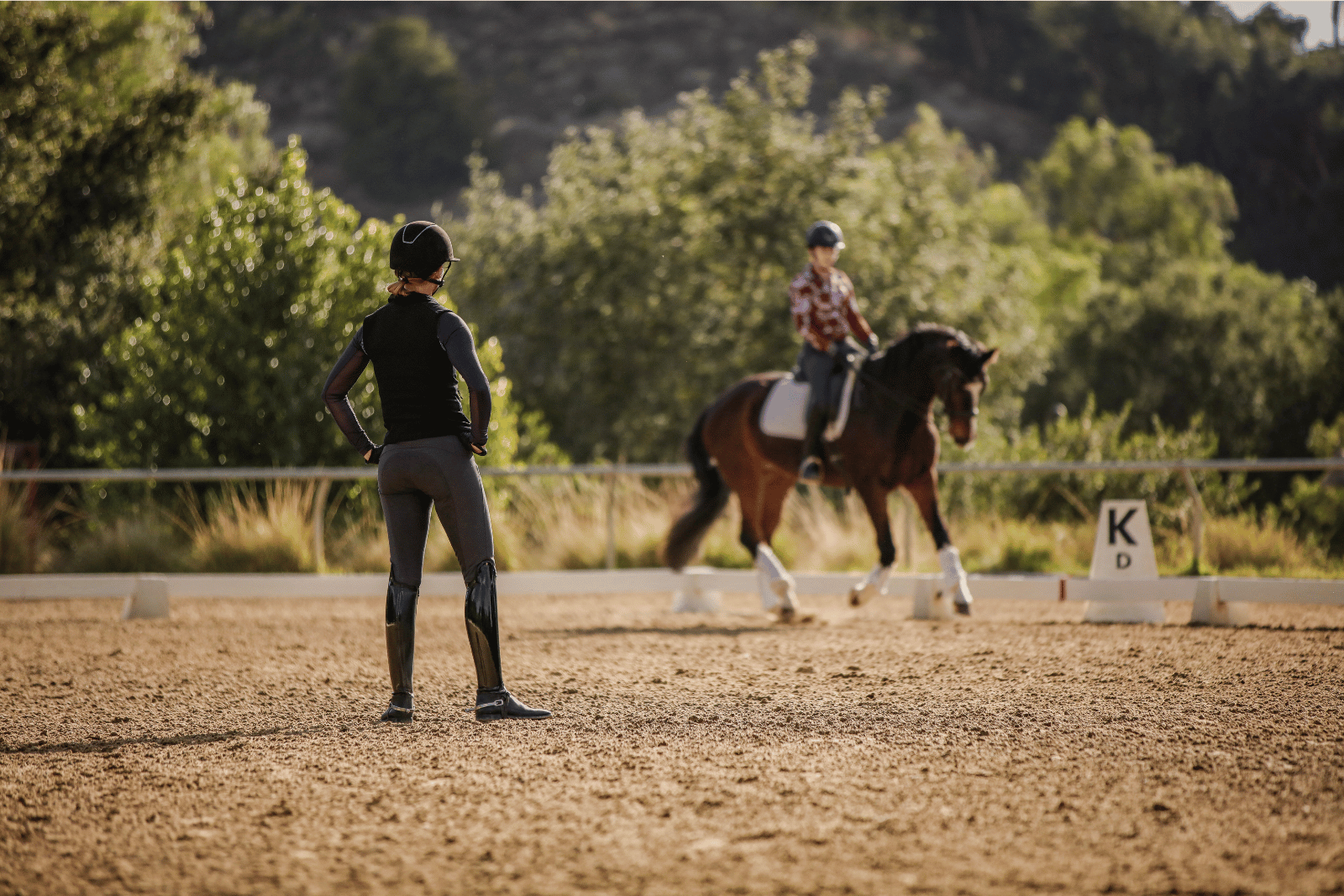 A person in riding gear stands on a sandy arena, watching another canter V1 around the ring. Trees and greenery fill the background, with the letter K sign visible—an inviting scene for any Open Page of equestrian life.
