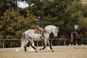 A person in a cowboy hat rides a gray horse at canter V1 in an outdoor sandy arena, smiling at the camera; another rider on a dark horse is visible in the background, surrounded by trees.
