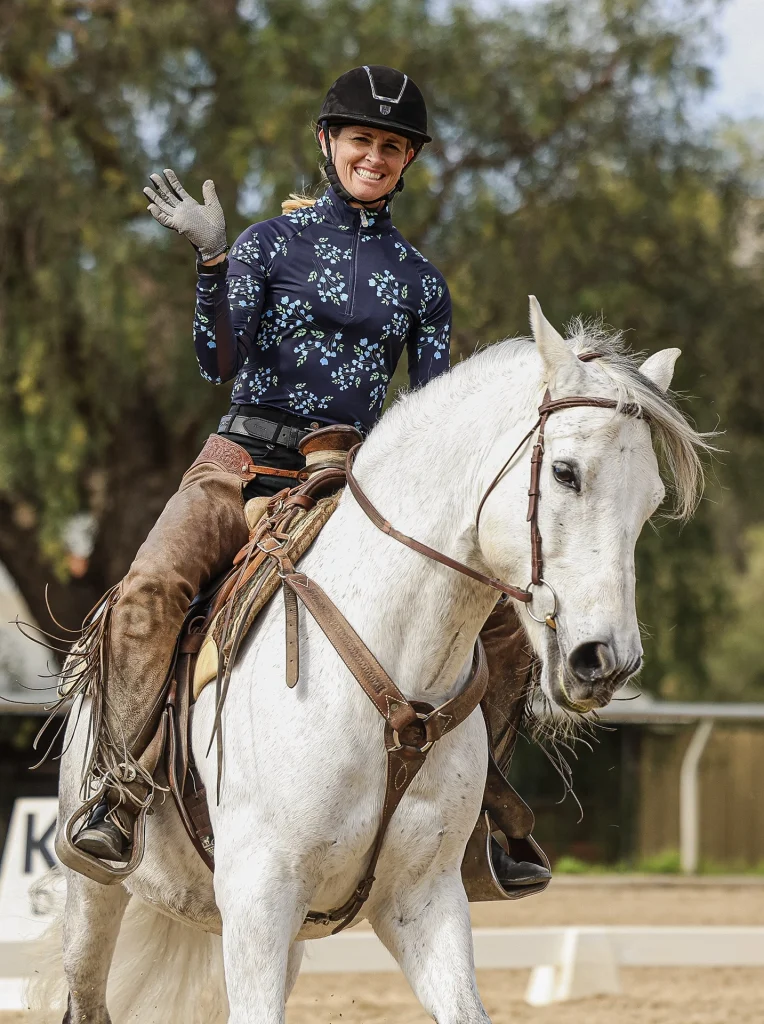 A smiling person in riding gear waves while sitting on a Canter V1 white horse outdoors. Trees and a fence are visible in the background, highlighting a joyful moment captured in an open page of adventure.