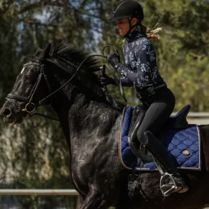 A person in equestrian gear rides a black horse with a blue Canter V2 saddle pad outdoors, set against a blurred background of trees.