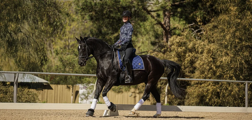 A person wearing riding gear and a helmet is cantering a dark horse with white markings in an outdoor sandy arena, surrounded by trees and a fence, enjoying the fresh air on this Open Page of their riding journey.